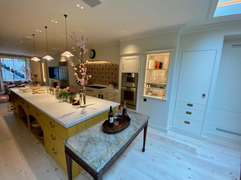 Kitchen with a long yellow island, stone worktop, built-in shelving, fitted cupboards and a patterned tiled splashback