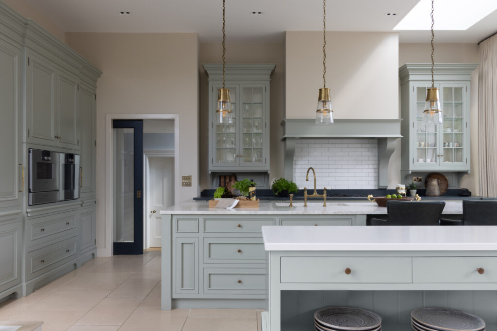 Sage Green bespoke kitchen with glazed display cabinets, brass pendant lights and a stone-topped central island