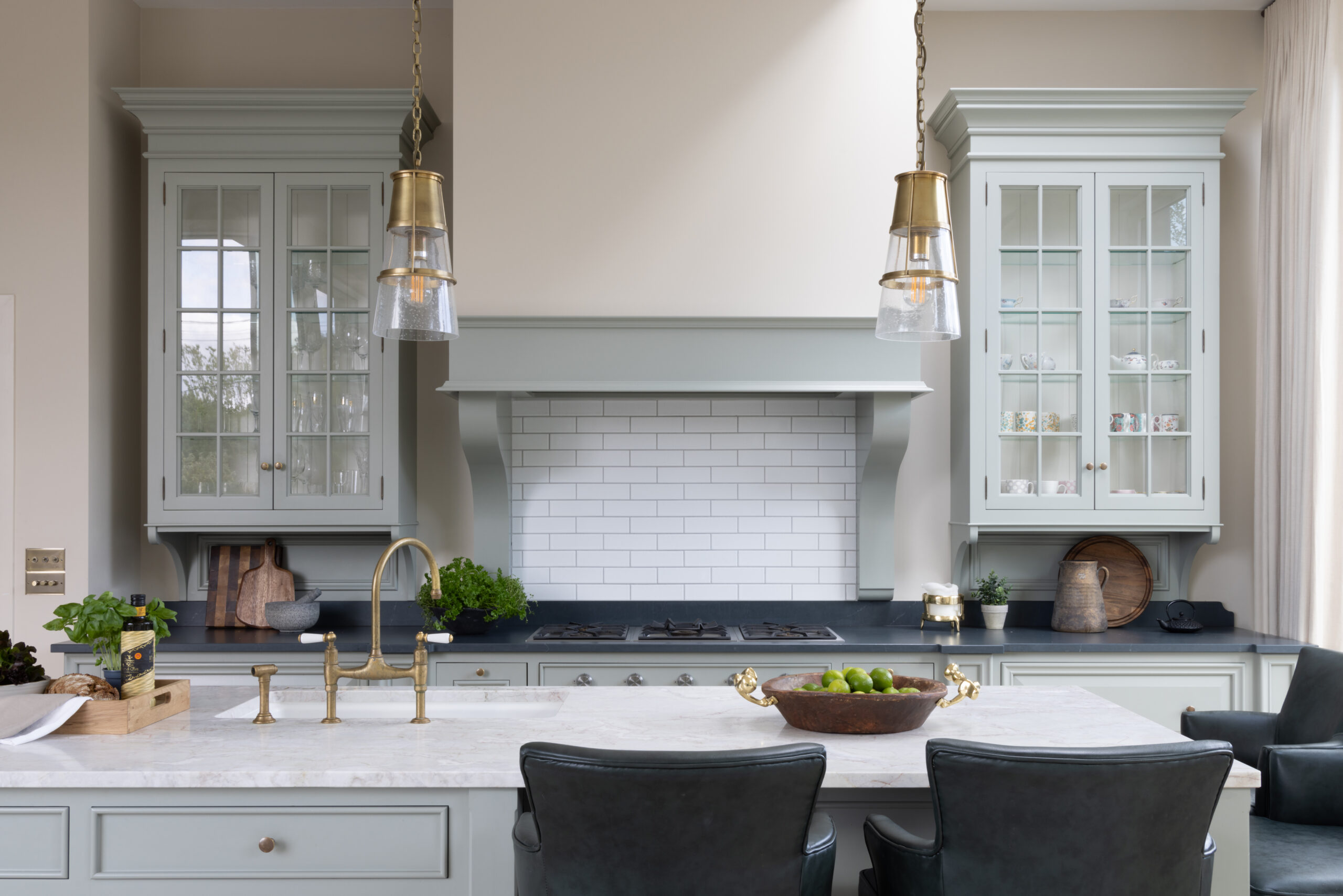 Sage green bespoke kitchen with glazed display cabinets, brass pendant lights and a central island with stone worktop