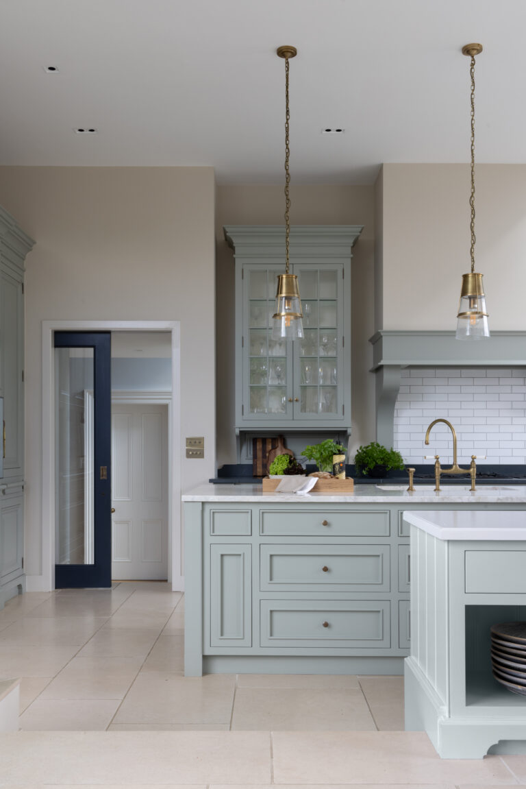Sage green kitchen island with brass pendant lights, glazed display cabinet and brass tap against metro tiled splashback