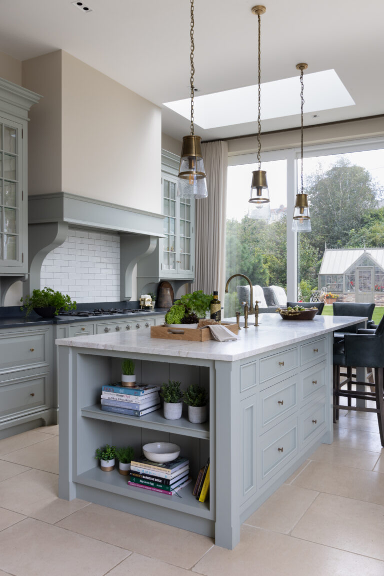 Sage Green bespoke kitchen island with stone worktop, open shelving and brass pendant lights beneath a skylight