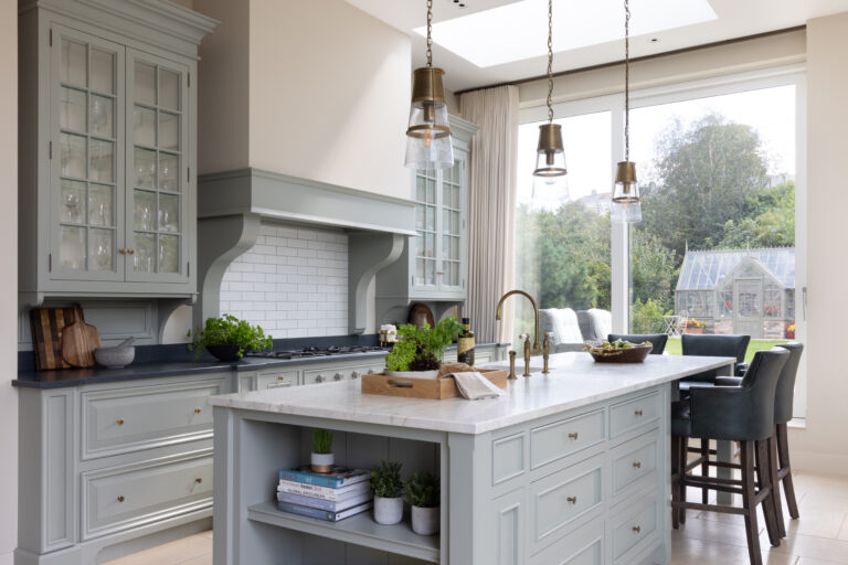 Sage cabinetry with glazed dressers and brass fittings surrounds a marble-topped island beneath tall windows