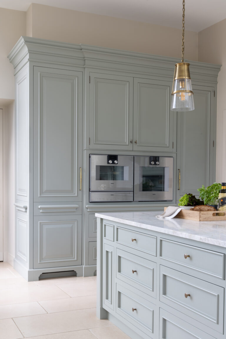 Sage-painted cabinetry with bolection mouldings, integrated ovens and a brass pendant light above the island