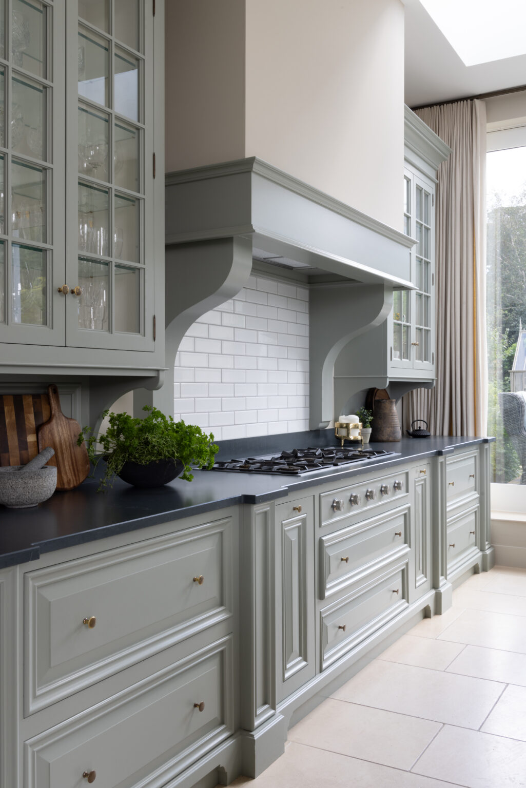 Sage Green bespoke kitchen with a mantled cooker hood, glazed cabinetry, dark worktop and gas range