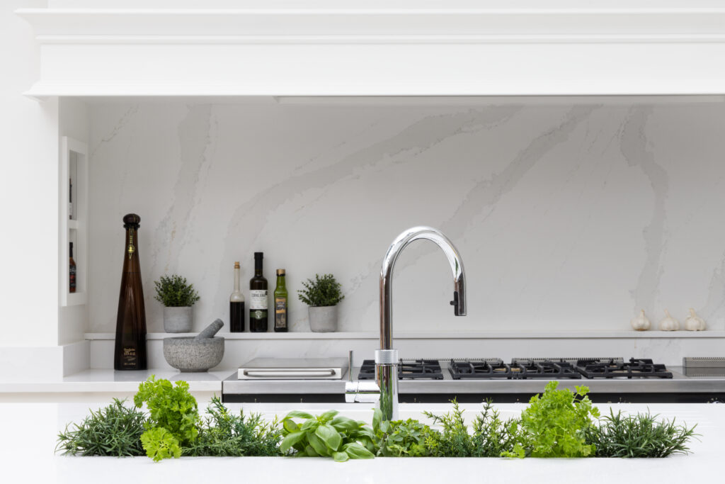 Kitchen sink filled with fresh herbs beneath a chrome tap, with gas hob and marbled splashback behind