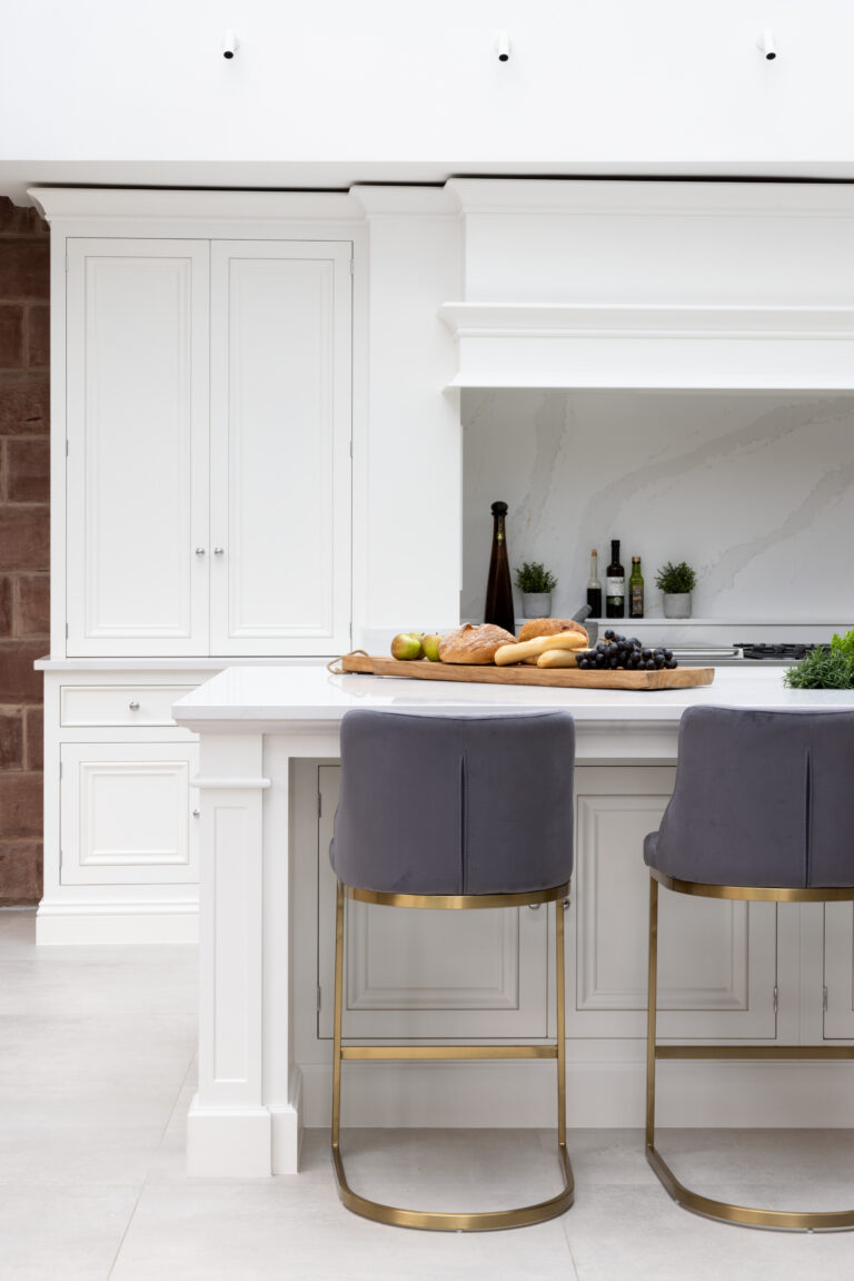 White bespoke kitchen island with grey velvet bar stools, brass legs, and an exposed brick wall alongside