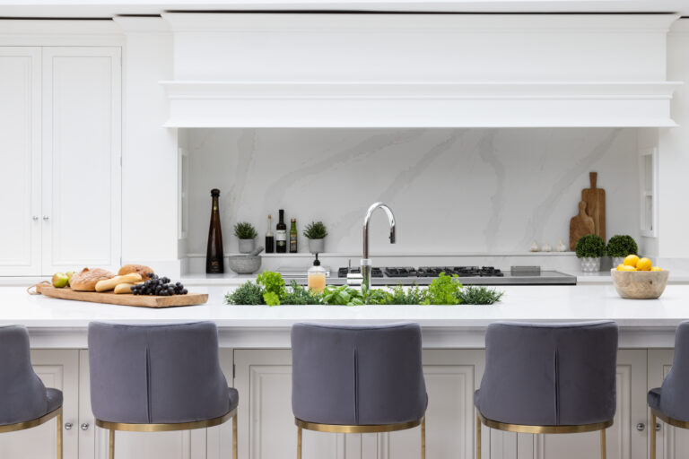 White kitchen island with stone splashback, grey velvet bar stools and fresh herbs arranged along the worktop