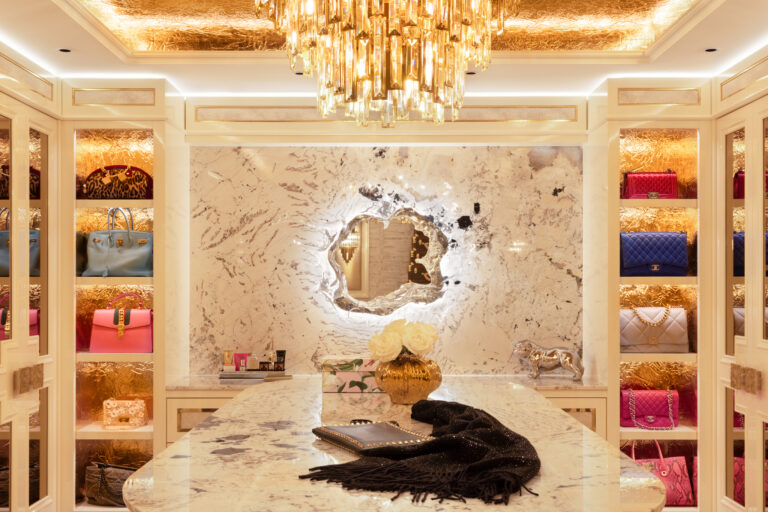 Dressing room with gold-foil lined shelving displaying designer handbags, a central island and sculpted mirror beneath a crystal chandelier