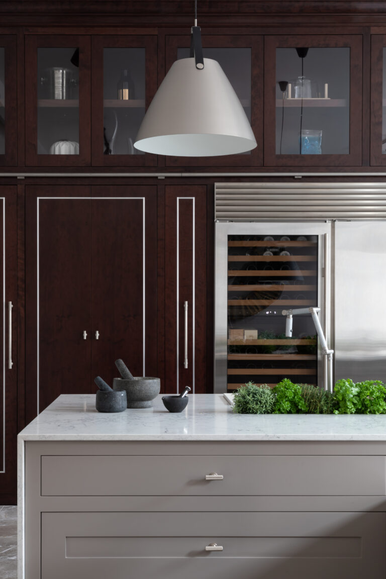 Kitchen island with pale stone worktop, pestle and mortars, fresh herbs, and cherry wood cabinetry with wine fridge behind
