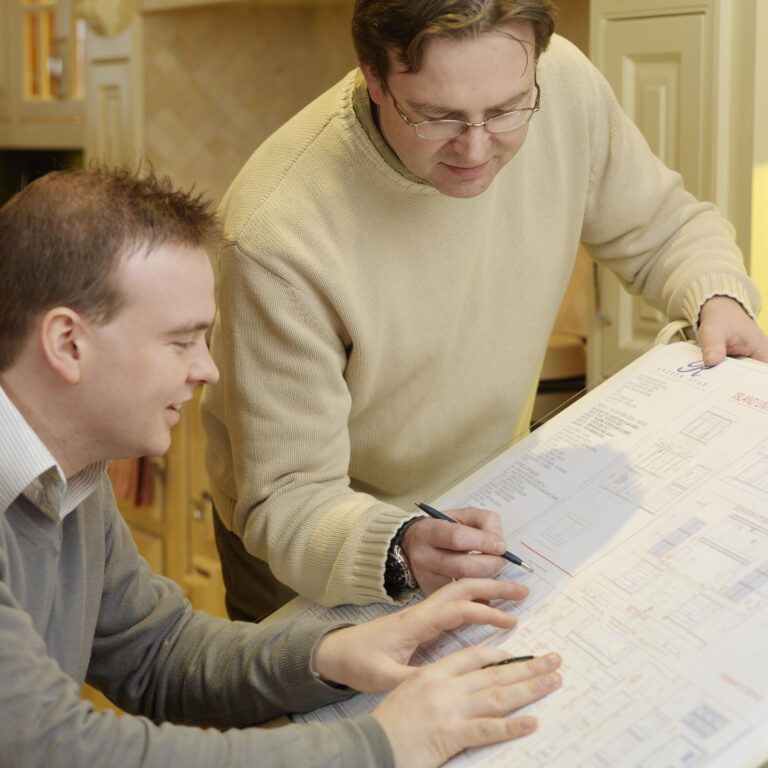 Two men review a detailed kitchen island design drawing together, with painted cabinetry visible in the background