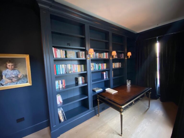 Study with floor-to-ceiling navy bookshelves, small wall lamps, a wooden desk and a framed portrait on the wall