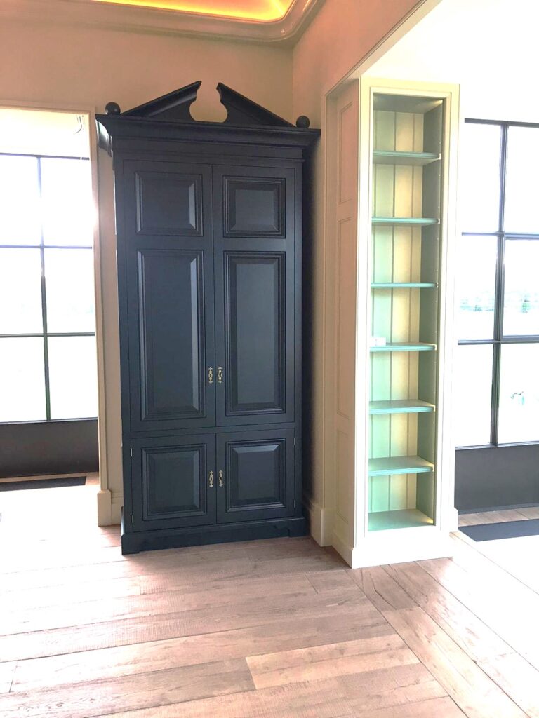 Tall panelled cupboard with a broken pediment top stands beside a slim built-in shelved alcove unit in a hallway