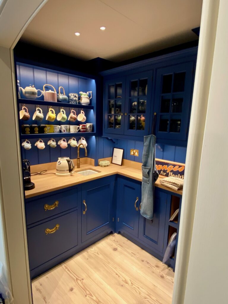 Small butler's pantry with dark blue cabinetry, brass handles, open shelving of teapots and mugs, and a wooden worktop with sink