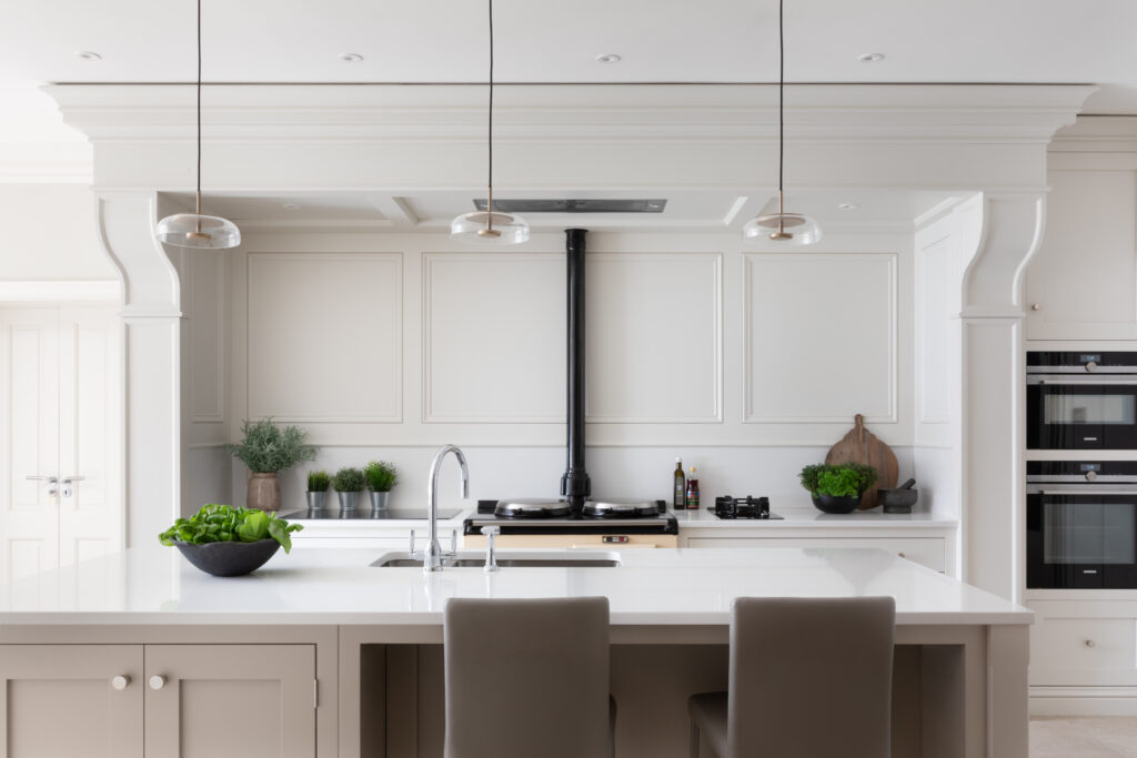 Kitchen island with pale cabinetry and white worktop facing panelled walls, pendant lights and an Aga with built-in ovens beyond