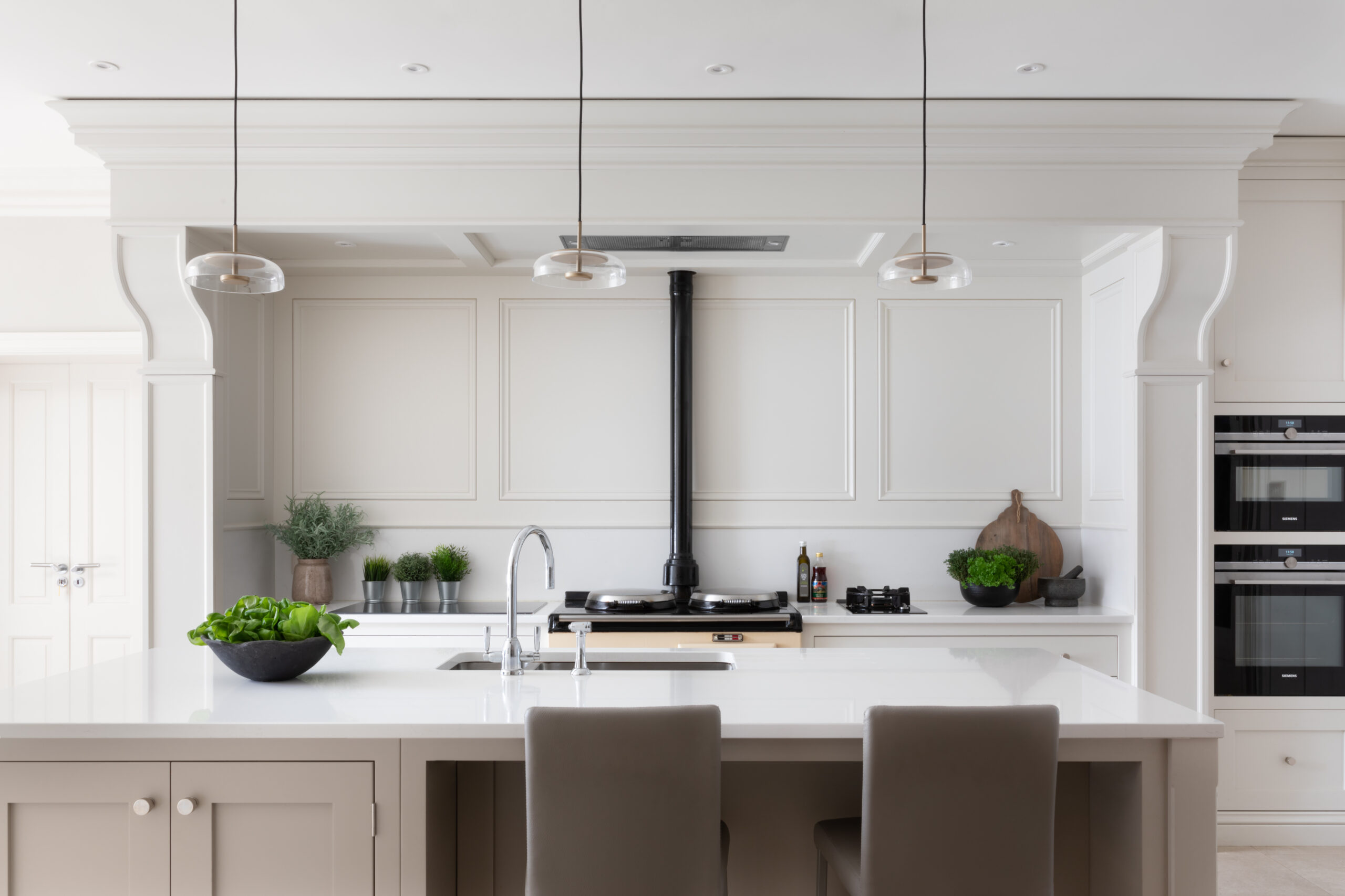 Kitchen island with pale cabinetry and white worktop facing panelled walls, pendant lights and an Aga with built-in ovens beyond