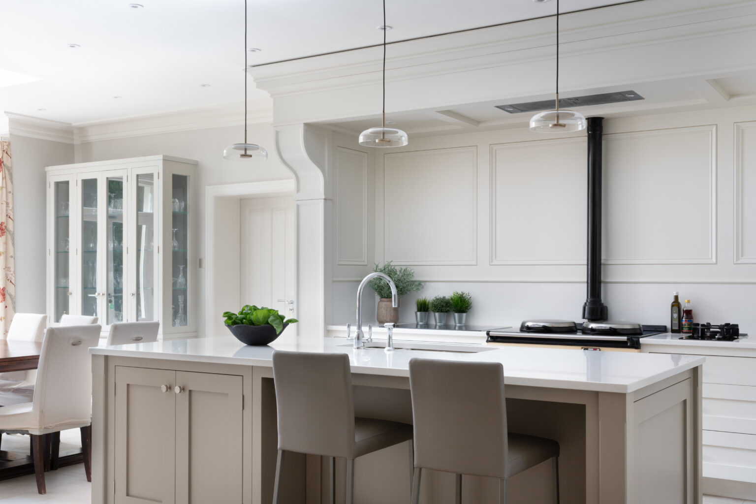 Kitchen island with pale cabinetry and bar stools, facing an Aga range with panelled walls and glazed display cabinet