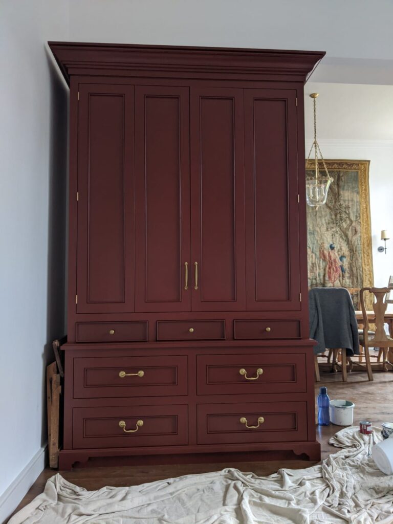 Tall painted cabinet with panelled doors, brass handles and multiple drawers, standing against a dining room wall