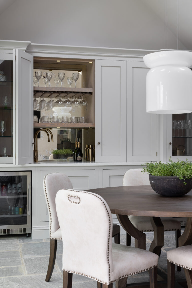 Bespoke home bar cabinet with glassware and brass tap, beside a round dining table with studded chairs
