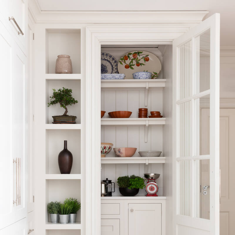 White glazed cupboard door opens onto a larder with painted shelving displaying crockery, bowls and plants