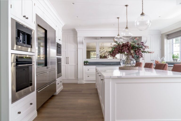 White bespoke kitchen with large island, integrated appliances, pendant lights and dining area with pink chairs