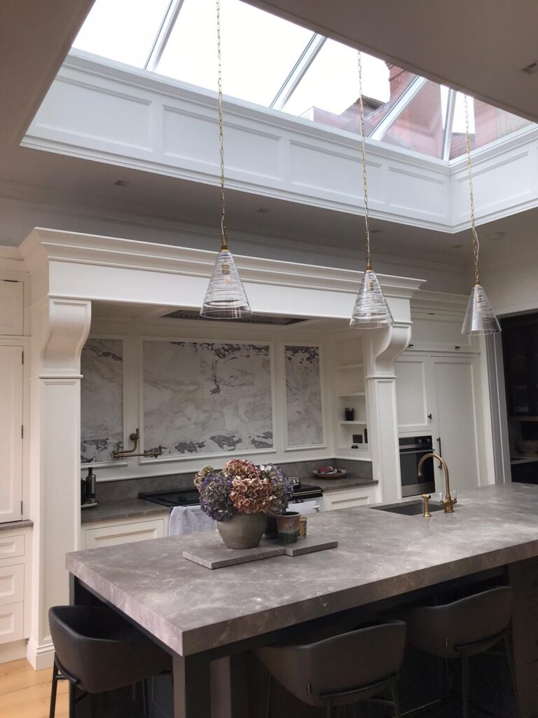 Bright kitchen with a large island, brass taps, glass pendant lights and a stone splashback beneath a skylight