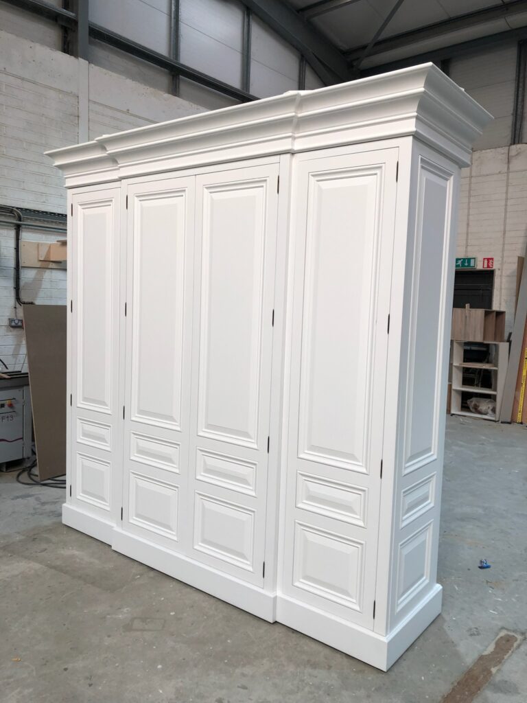 A large white panelled wardrobe with a moulded cornice top stands finished in the workshop before delivery