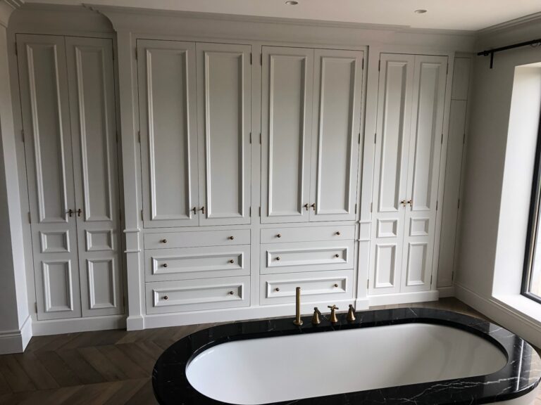Bathroom with painted panelled wardrobes and drawers, brass handles, beside a freestanding bath with stone surround