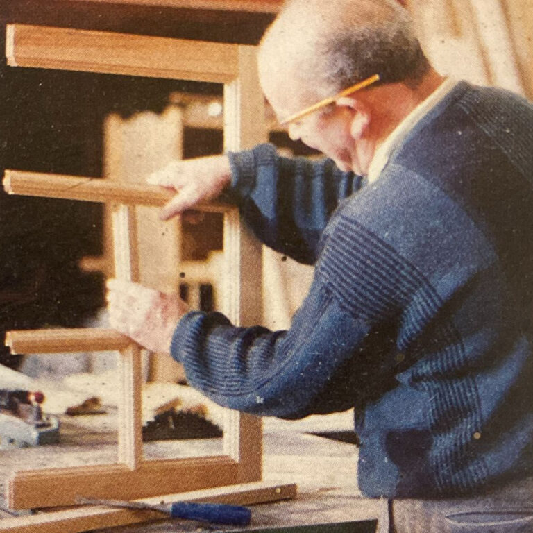 A craftsman in a workshop examines a timber frame joint, pencil behind his ear, checking the joinery closely