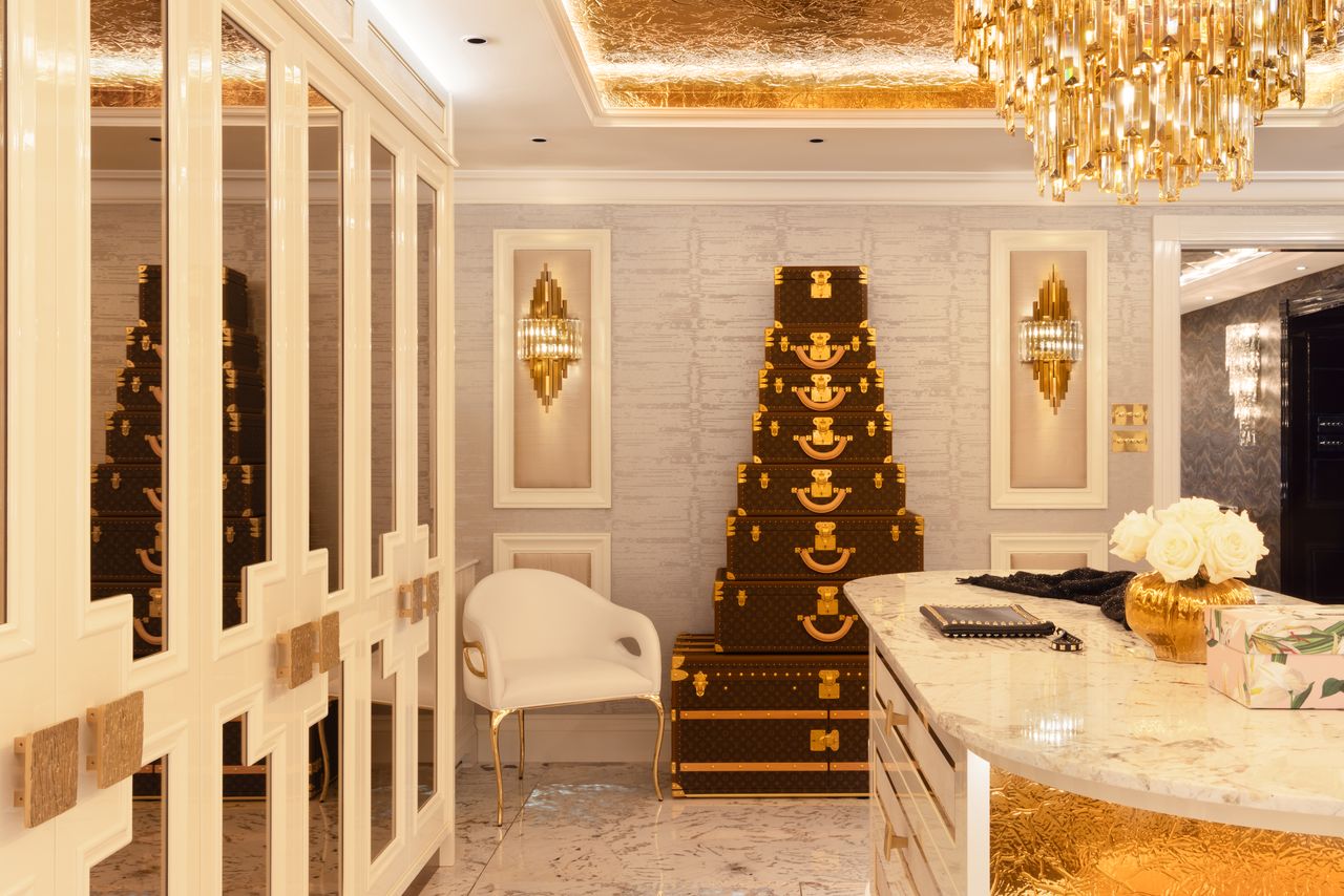 Dressing room with mirrored wardrobes, gilt ceiling, crystal sconces and a stacked trunk display beside a stone-topped island