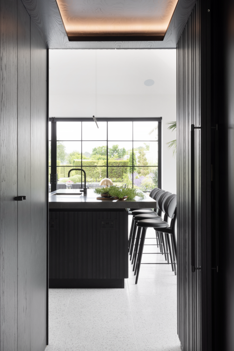 Doorway view into the kitchen island with bar stools, framed by Crittall glazing overlooking the gardens