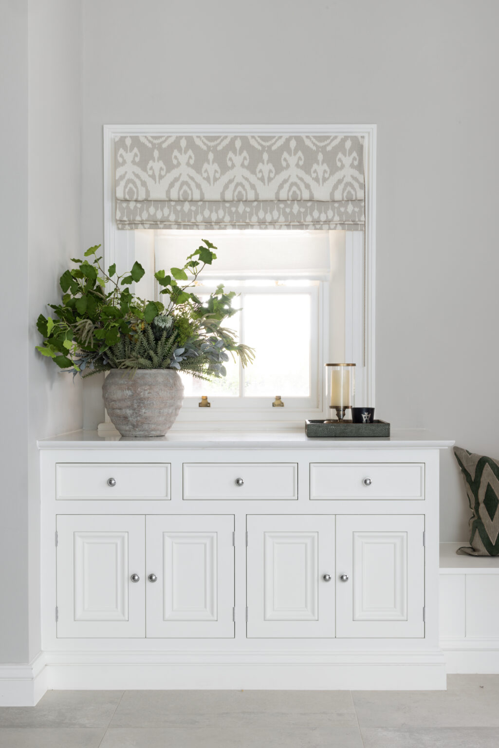 White built-in cabinetry beneath a window, topped with a foliage-filled vase and patterned roman blind above