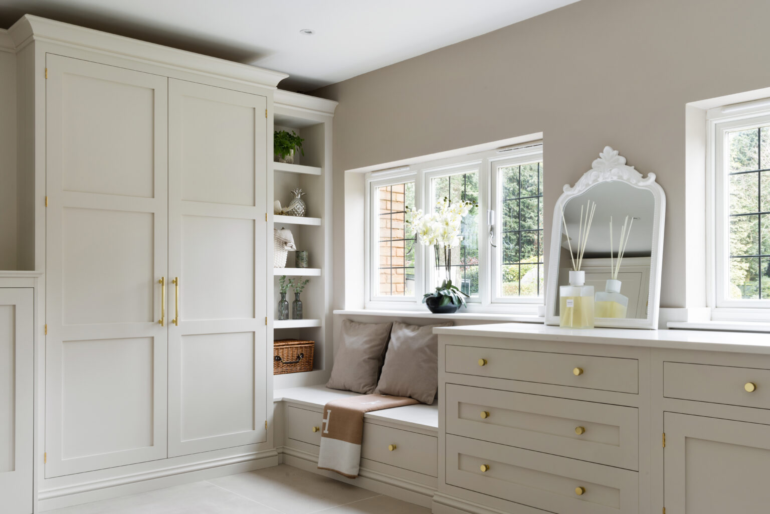 Fitted boot room in Little Greene 'Joanna' with floor-to-ceiling cupboards, open shelving, a cushioned window seat and brushed gold handles