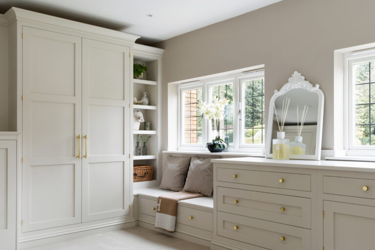 Fitted bedroom joinery with tall wardrobe, open shelving, window seat with cushions, and chest of drawers with brass handles