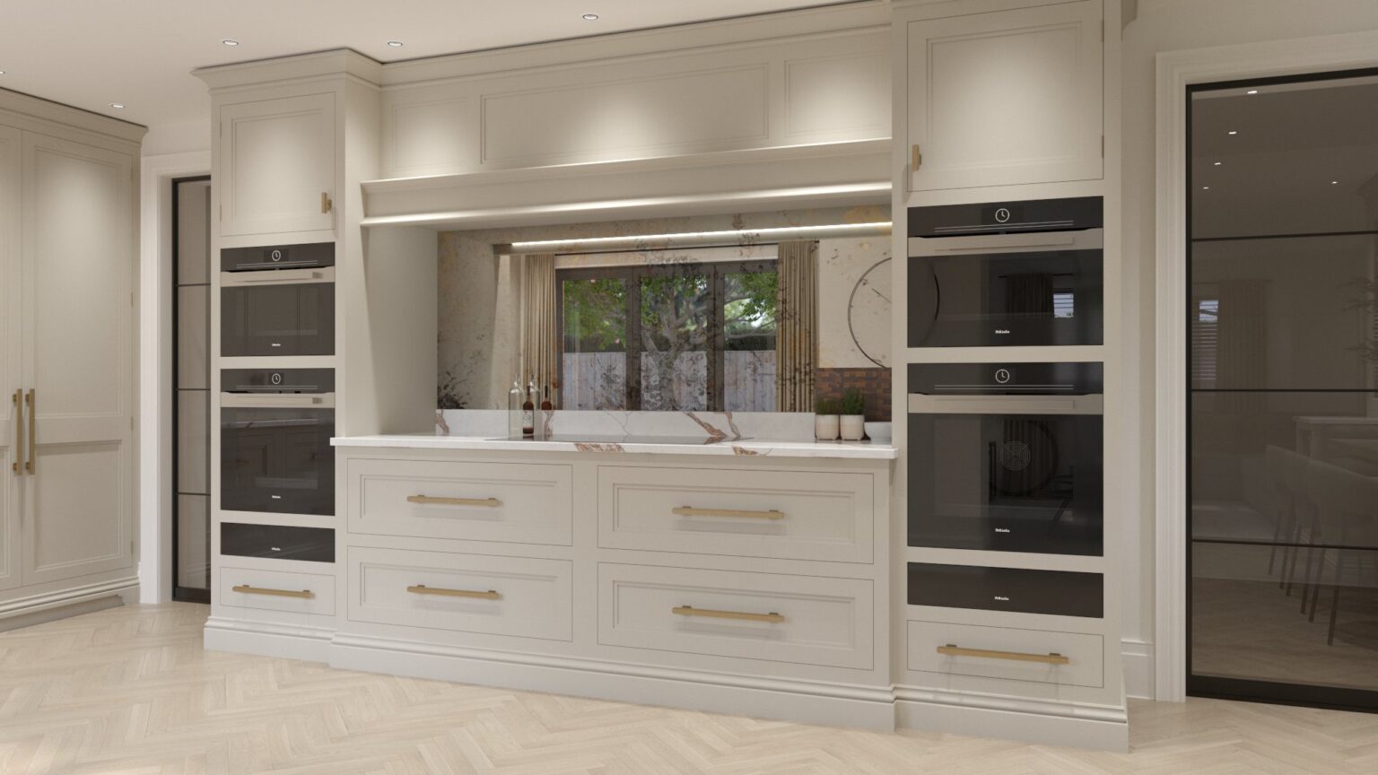 Kitchen wall of cream cabinetry with integrated ovens, brass handles and a stone-topped worktop beneath a mirrored splashback