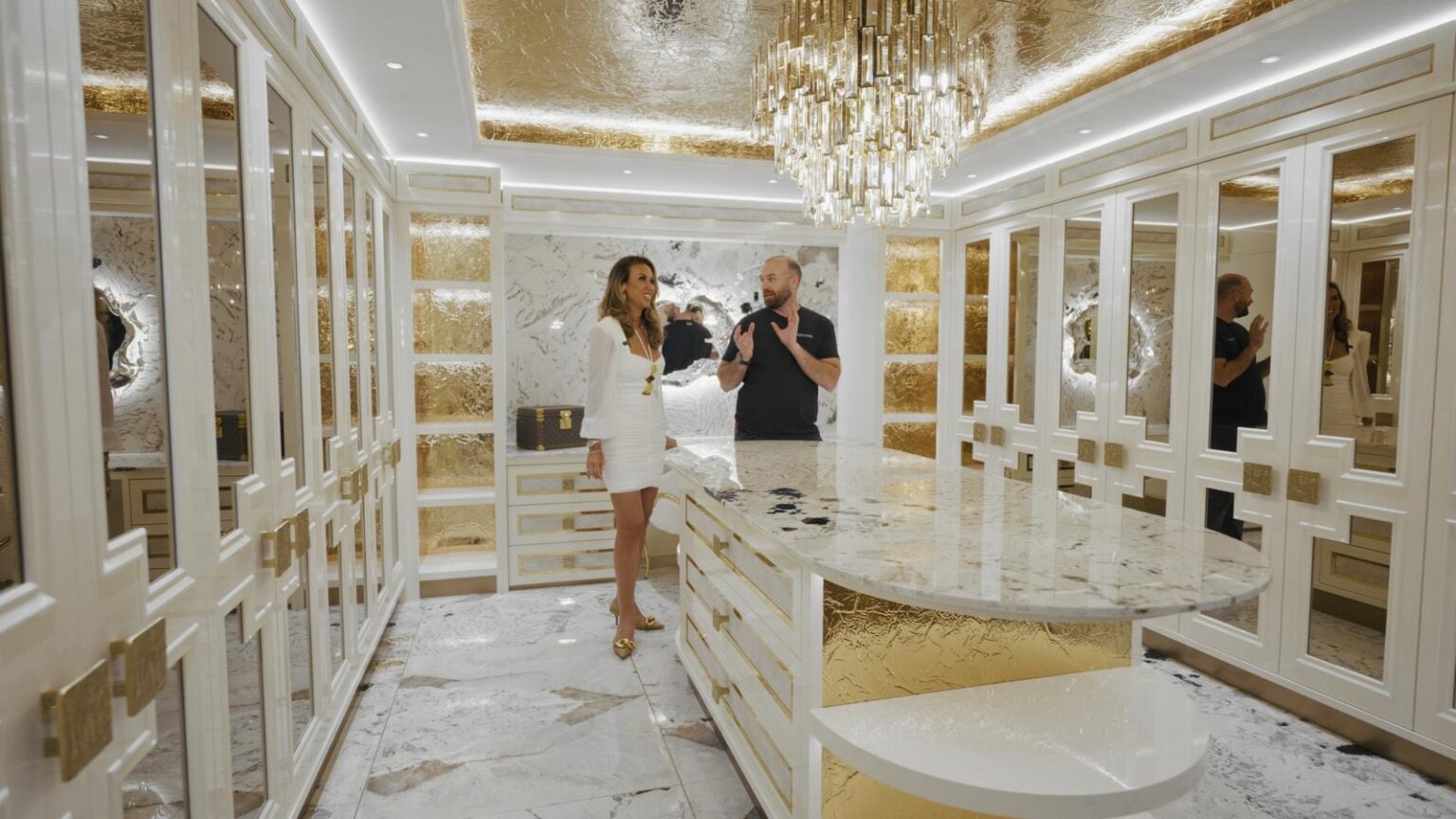 Curved ivory onyx island beneath a gold leaf ceiling, surrounded by high-gloss ivory cabinetry and mirrored wardrobes