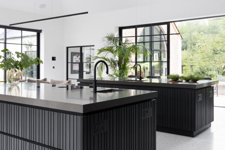 Twin slatted ash islands with dark worktops face Crittall glazing, framed by tall windows and greenery