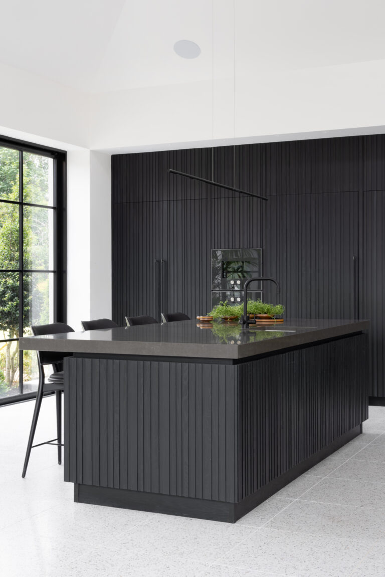 Kitchen island with slatted dark cabinetry and stone worktop, set beside Crittall glazing overlooking the garden