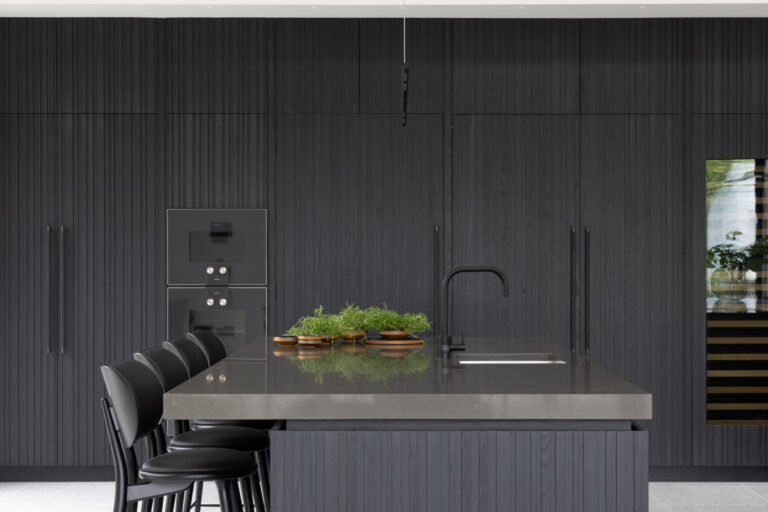 Dark slatted ash cabinetry wall with integrated Gaggenau appliances, a stone-topped island, sink, tap and bar stools