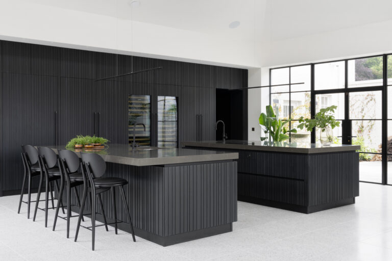 Two slatted ash islands with bar stools face Crittall glazing, grey worktops and integrated appliances along the dark cabinetry wall