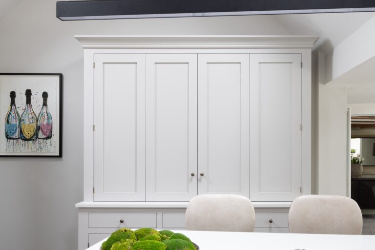 Tall white panelled cupboard with bi-fold doors concealing the wet bar, set above drawers and a bench seat