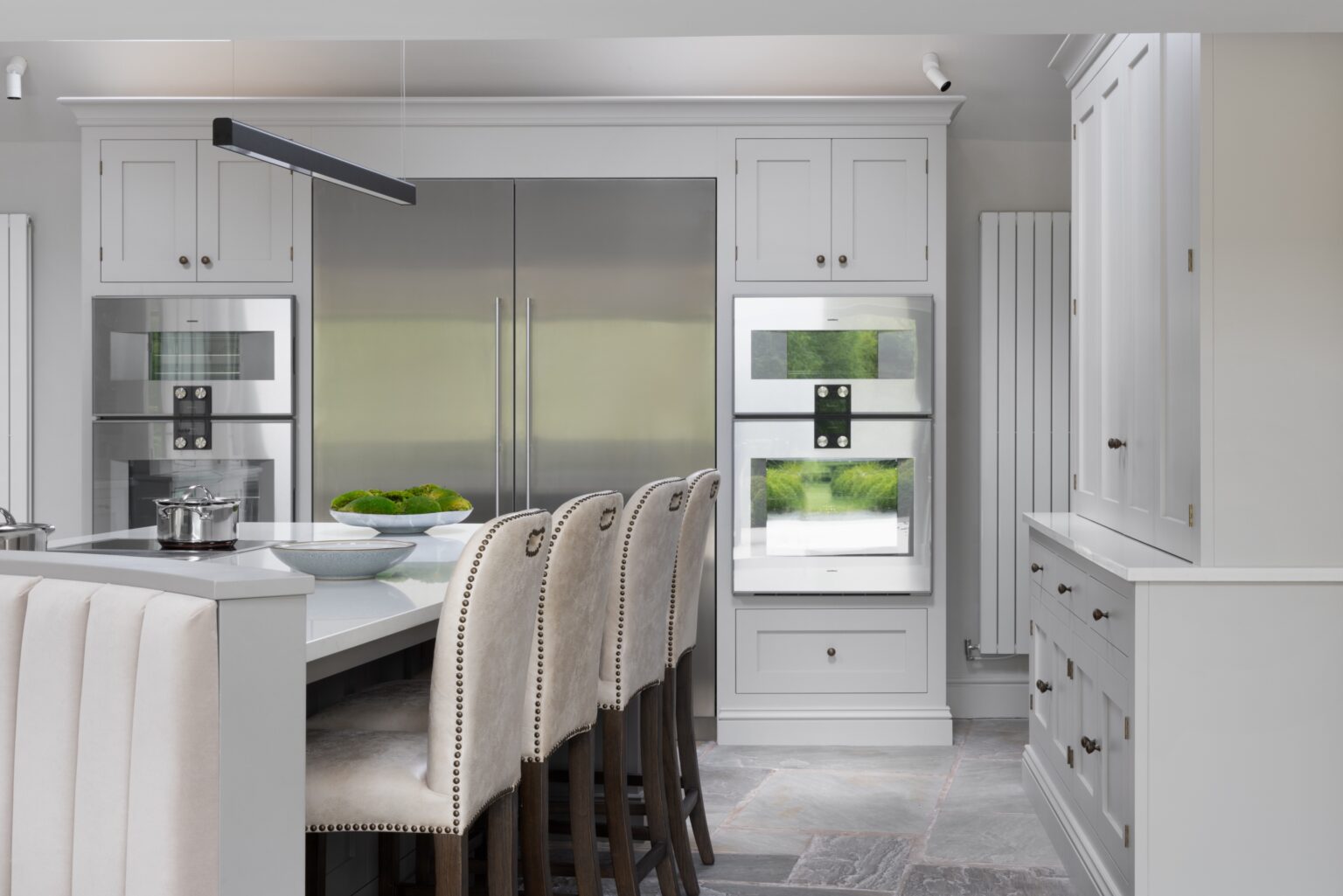 Kitchen island with upholstered stools beside a large fridge freezer and built-in double ovens flanked by shaker cabinetry