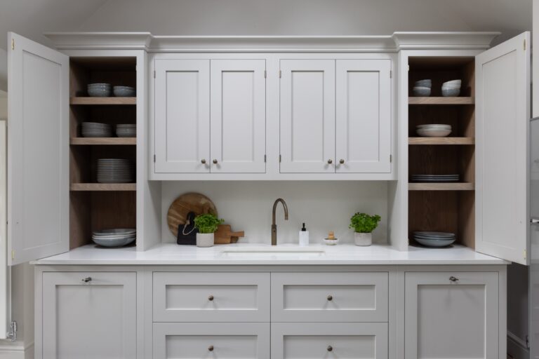 Sink unit flanked by open shelved cupboards displaying crockery, with a brass tap and bowl set into a white worktop