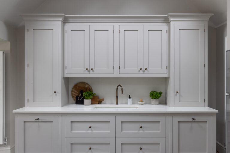 Shaker-style wall and base cabinetry framing a sink with brushed tap, set into a white stone worktop