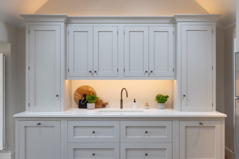 Fitted grey Shaker cabinetry frames a sink set into a white worktop, lit by under-cabinet lighting