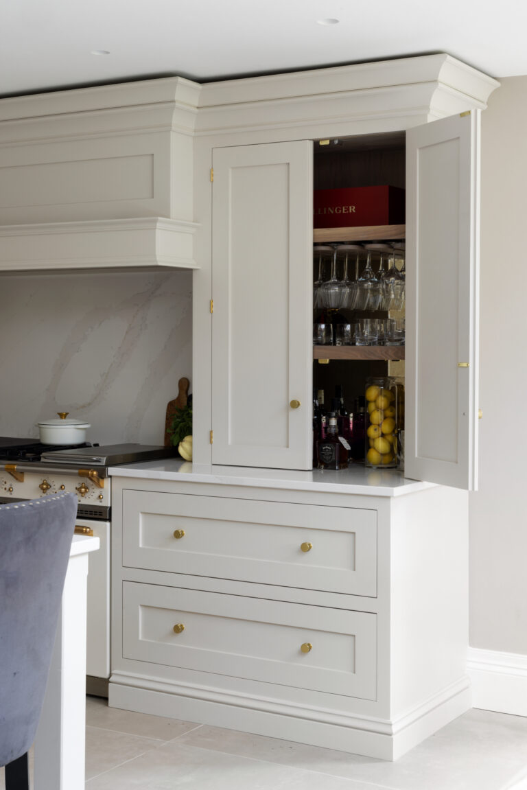 Open drinks cabinet with brass handles revealing glassware and bottles, beside the range cooker and stone splashback