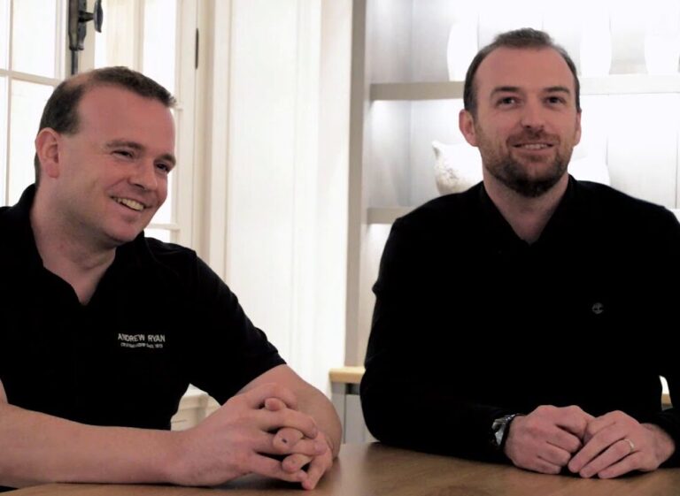 Two men seated at a wooden table, smiling, with a bright shelved display unit visible in the background