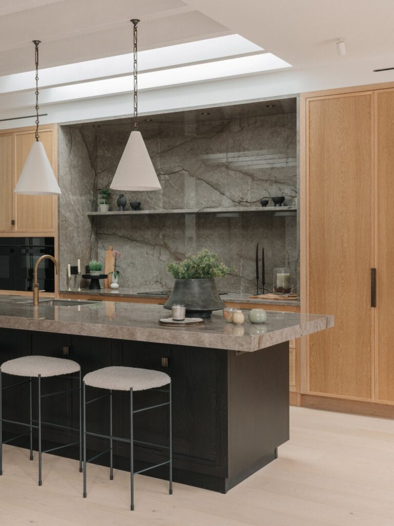 Black-stained oak island with a 70 mm Astana Grey stone top and stools, in front of a stone-lined hob wall with a floating stone shelf and tall Cotton White oak cupboards