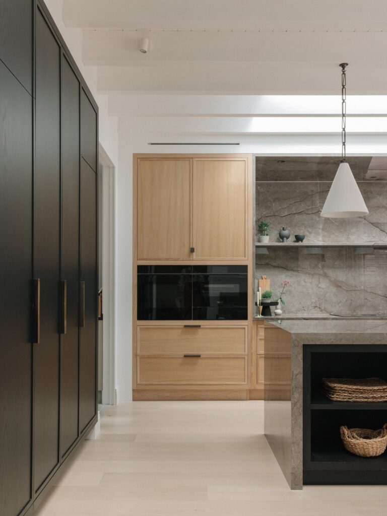 Tall black-stained oak cupboards beside a Cotton White oak oven housing, with the stone-topped island and its open shelf in front