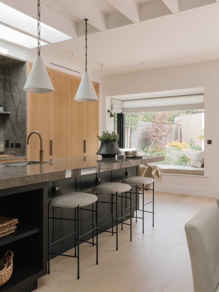 Black-stained oak island with upholstered stools under white cone pendants, tall Cotton White oak cupboards behind and a window seat overlooking the garden